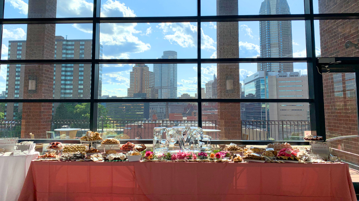 A long cookie table is set inside the Dougherty Ballroom, with floor-to-ceiling windows overlooking a city skyline. The table is covered with a pink tablecloth and filled with trays of assorted sweets and finger foods, including cookies, pastries, and small bites arranged on platters. In the center, an ice sculpture spells out the word “LOVE,” surrounded by colorful flowers. Brick columns frame the large windows, and tall buildings and blue sky with scattered clouds are visible outside, creating an airy, celebratory atmosphere.