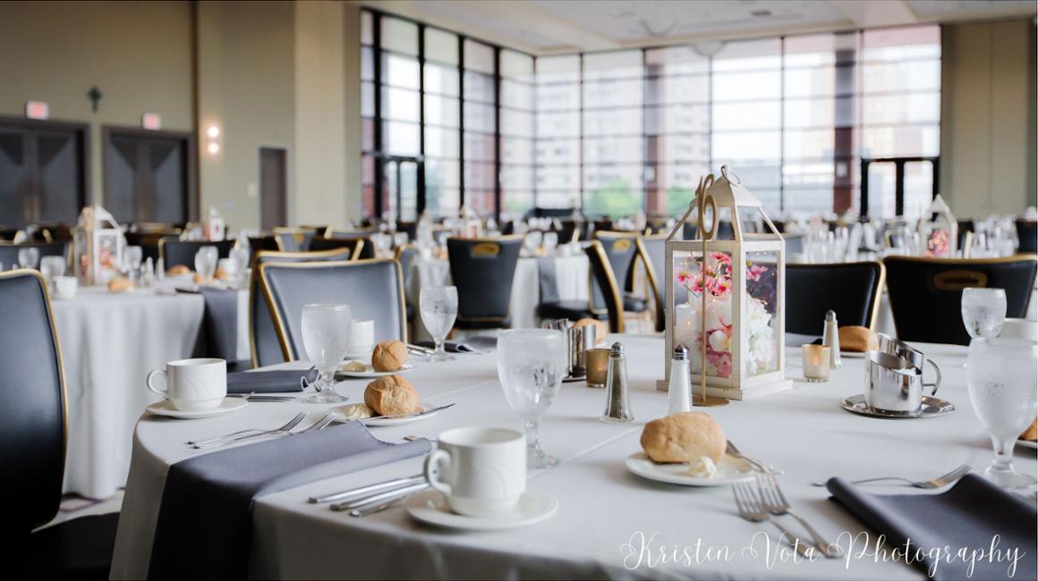 The Dougherty Ballroom, a bright banquet room, is set for a formal meal with multiple round tables covered in white tablecloths. Each place setting includes a white cup and saucer, water glass, silverware, folded gray napkin, and a small bread roll on a plate. The chairs are dark with gold trim. A decorative lantern centerpiece filled with pink and white flowers sits in the foreground on one table. Large floor-to-ceiling windows along the back wall allow natural light to fill the space, and additional similarly arranged tables extend throughout the room, ready for guests.