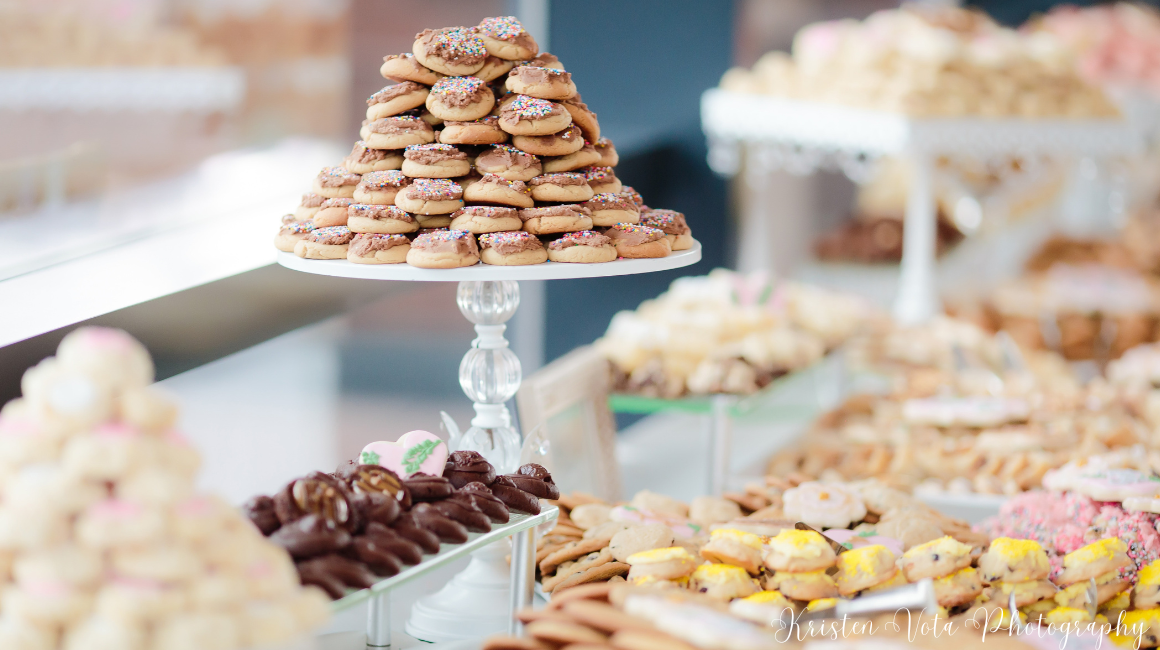 A cookie table display features multiple tiers of cookies arranged on white cake stands and trays. In the center, a pyramid-shaped stack of frosted cookies topped with colorful sprinkles sits on a raised pedestal. Surrounding it are assorted treats, including chocolate-dipped cookies, sandwich cookies with yellow cream filling, and various iced and decorated cookies in different shapes and colors.