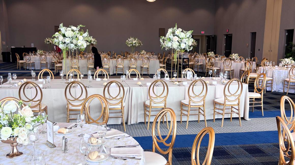 The Dougherty Ballroom is arranged for a formal event with long rectangular and round tables covered in light-colored tablecloths. Each place setting includes glassware, silverware, folded napkins, and a small bread roll. Gold circular-back rented chairs surround the tables. Tall floral centerpieces with white flowers and greenery stand on slender gold stands above several tables.