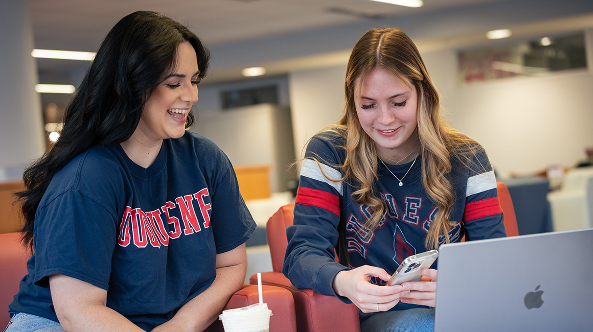 Students sitting in front of a laptop and looking at a smart phone