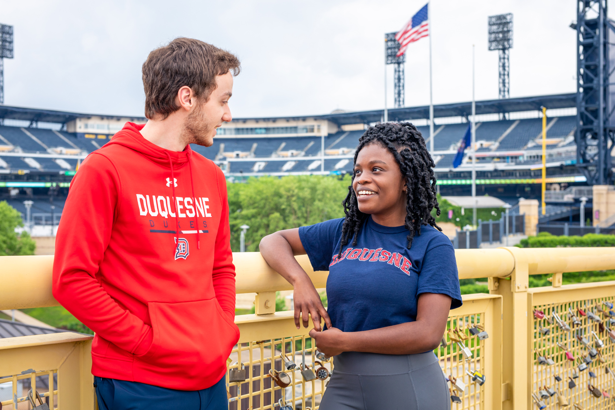 Female and male student talking on Pittsburgh bridge with PNC Park in the background.