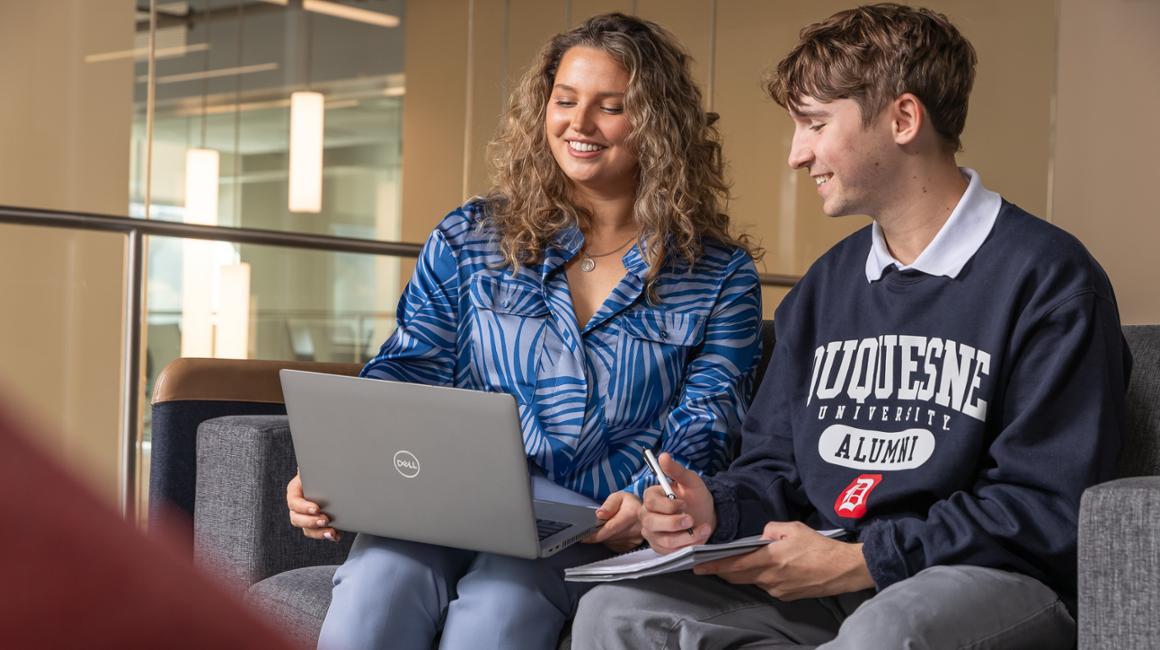 Two graduate students sit in a lounge and work together on a laptop.