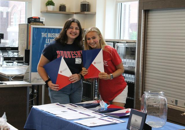 Two School of Business student ambassadors pose with red, white, and blue event folders. 