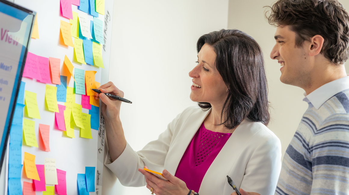 A professor and a student point to sticky notes on a whiteboard.