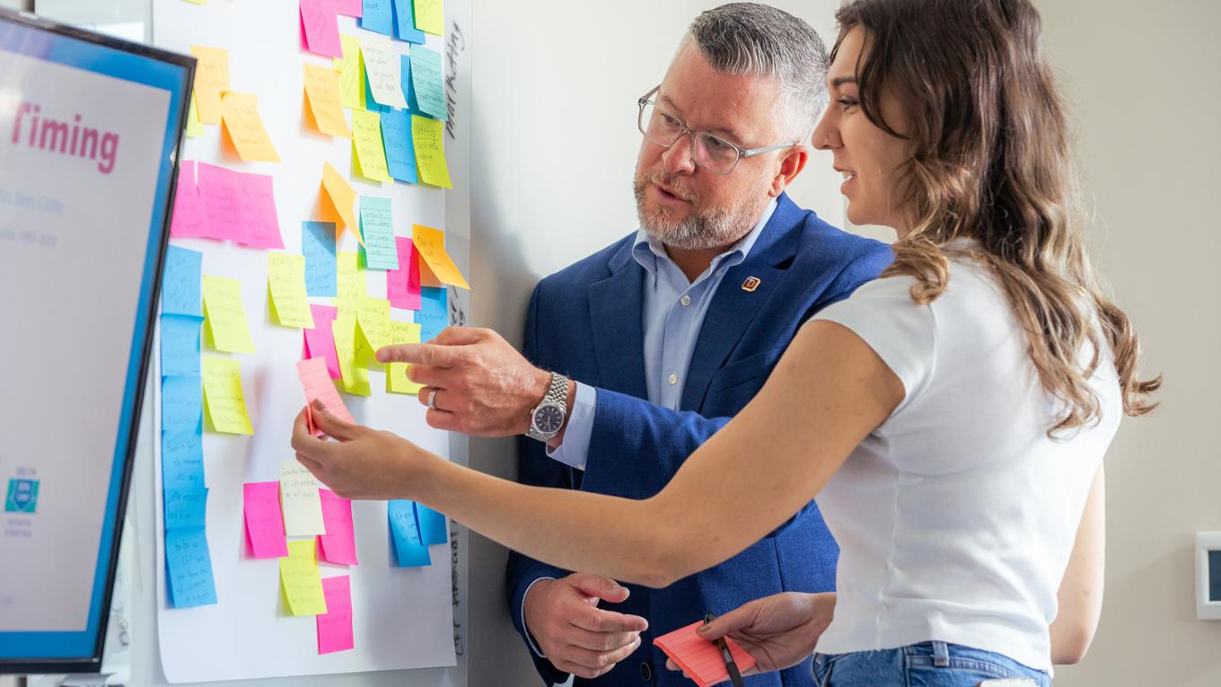 A professor and a student point to sticky notes on a whiteboard.