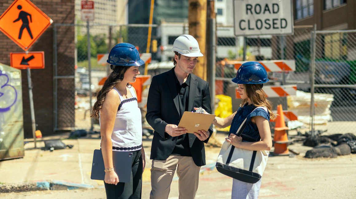 Business students wear hardhats and review plans at a construction site.