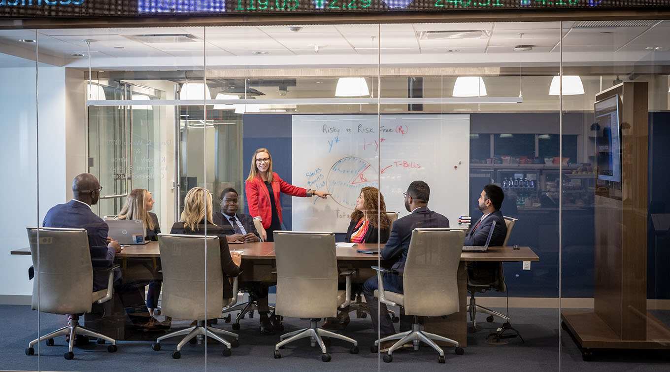 A wide photo of business students meeting in a glass-walled conference room. 