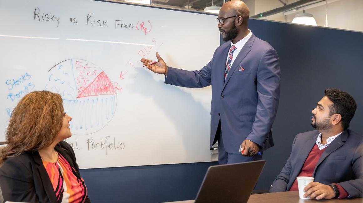 A man in a suit writing on a whiteboard with students.