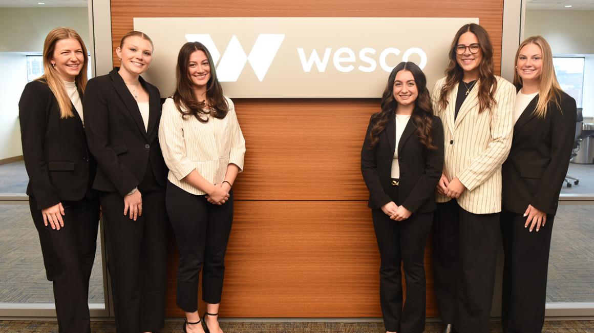Female business students in professional attire pose in front of a conference room at Wesco headquarters. 