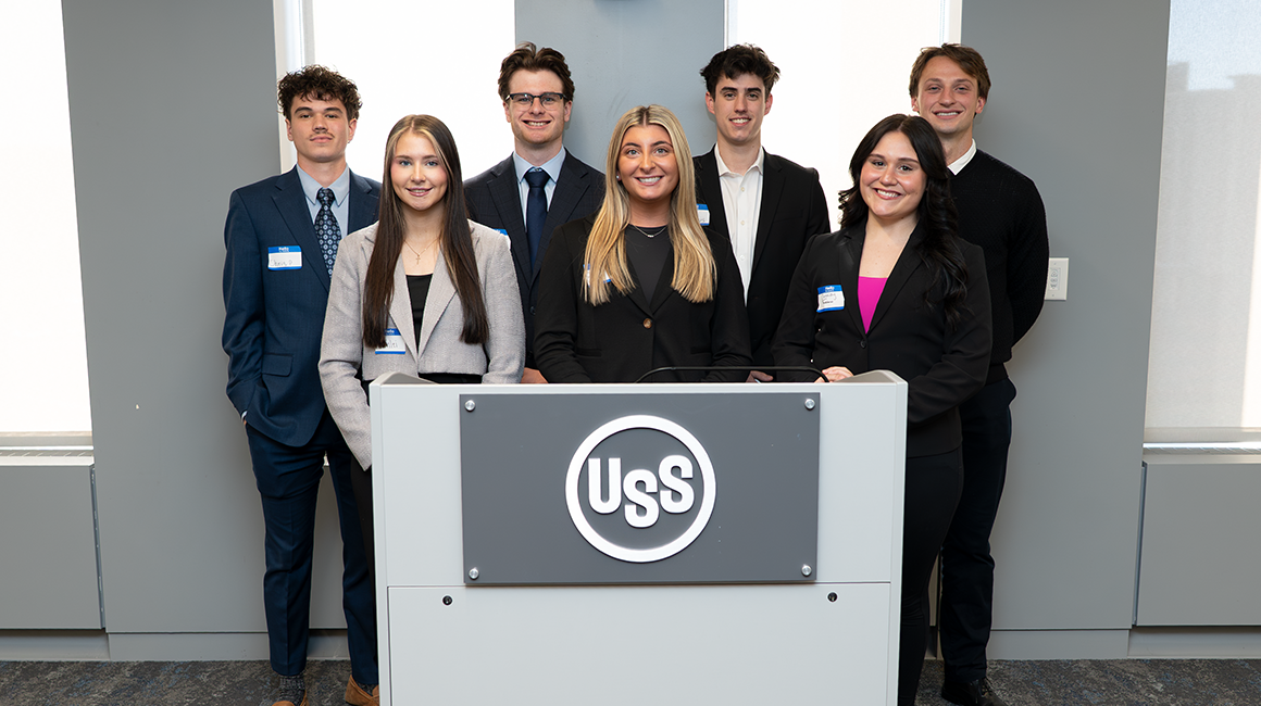 Business students in professional attire pose in front of a podium with a US Steel logo.