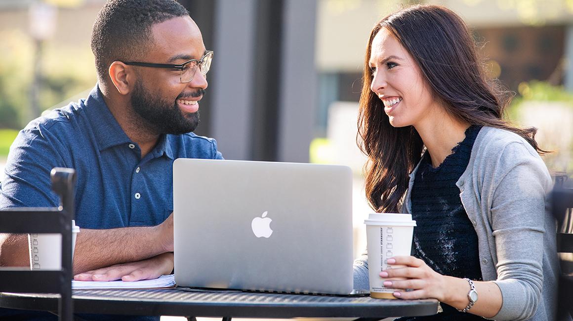 Two business students talk outside while sitting in front of a laptop.