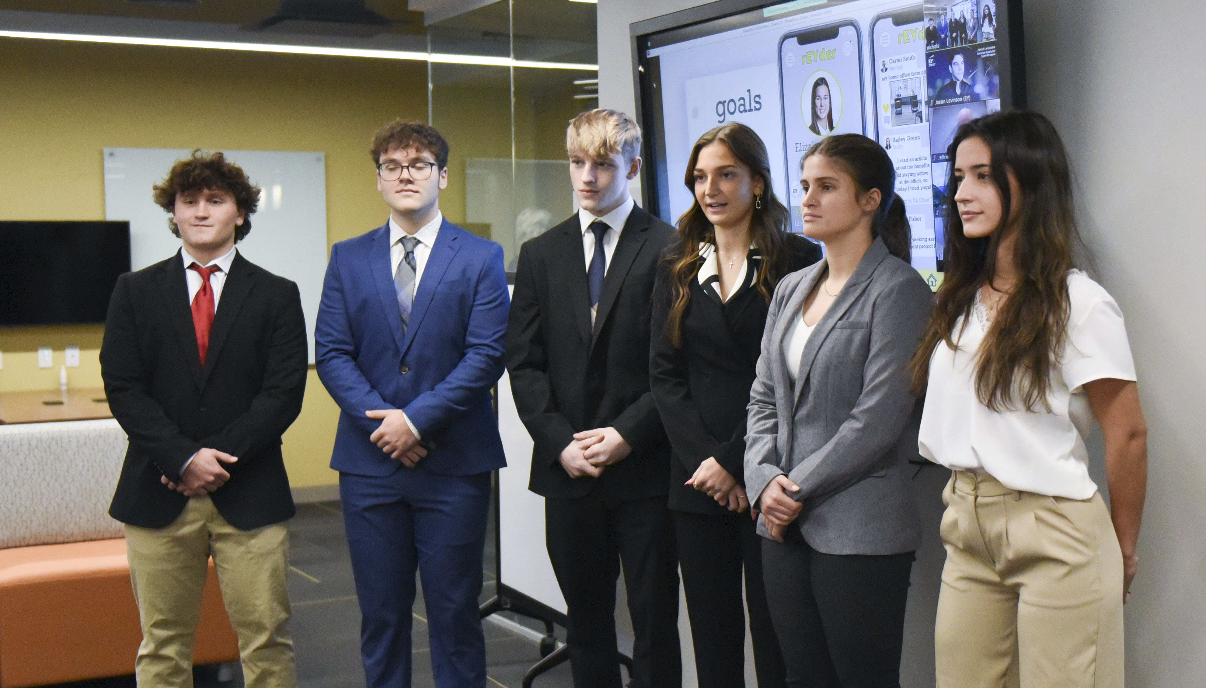 Six students in business attire stand in a row in front of a screen. 
