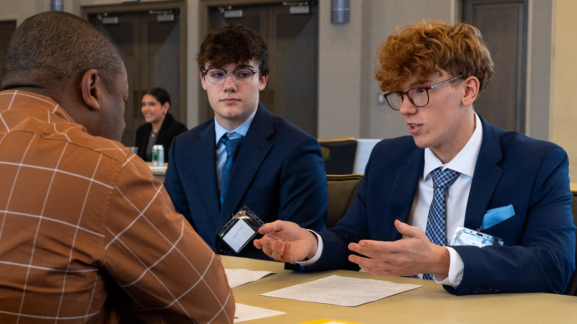 Two students sit facing a business professional. One of the students is expressive with his hands as he talks.