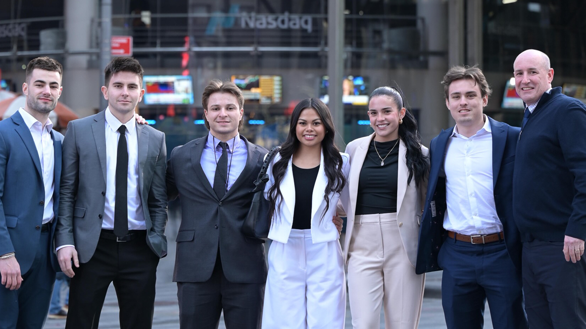 A team of students and a professor pose in front of the Nasdaq sign in Times Square in NYC.