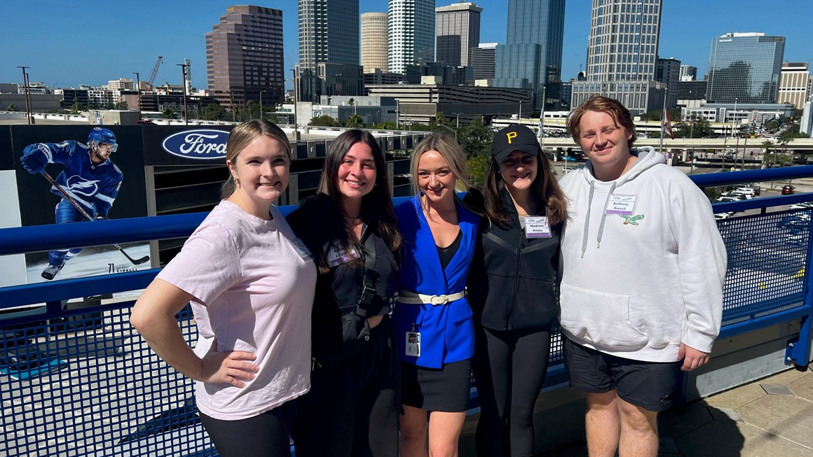A group of students pose outside in front of the Tampa city skyline. Tampa Bay Lightning signage can be seen in the background.