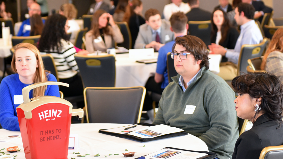 Students and professionals listen attentively at a networking event. A Heinz ketchup dispenser sits on the table.