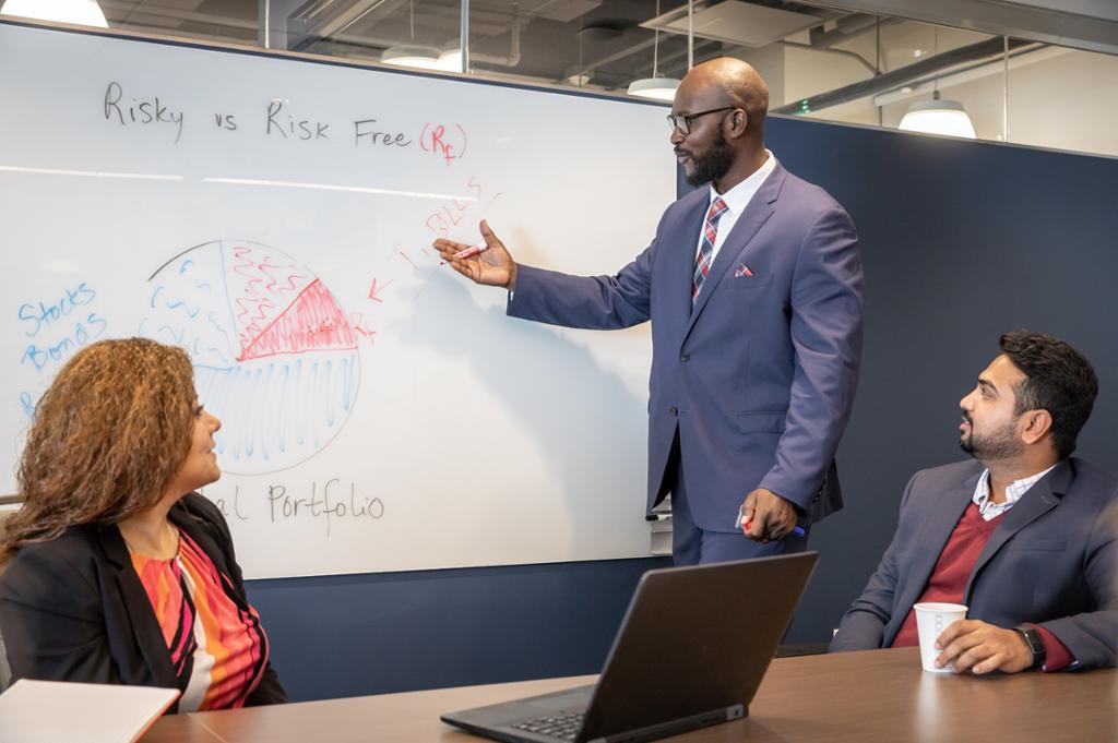A man in a suit writing on a whiteboard with students.