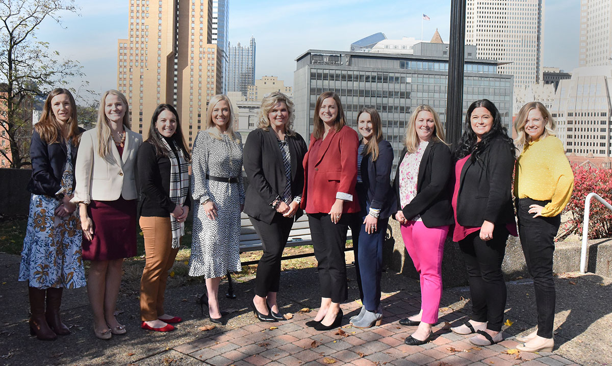 Ten women stand in a row outside with downtown Pittsburgh behind them. 