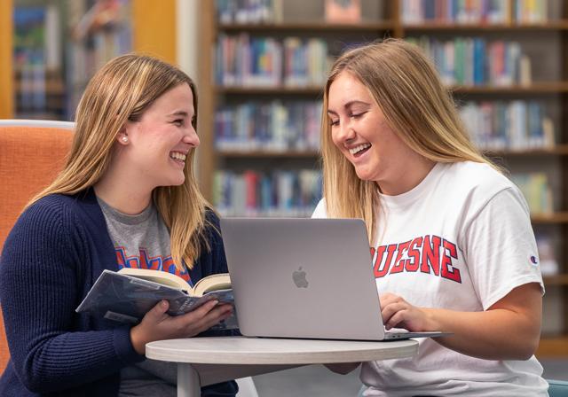 Two undergraduate scholarship students in library smiling and studying together.