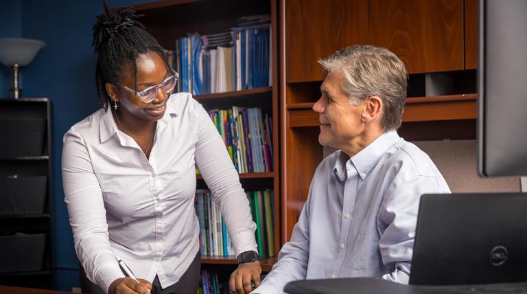 Photo of Duquesne faculty and student having a discussion at desk with bookshelf in background