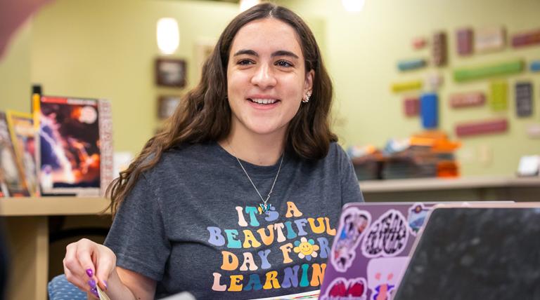 School of Education student wearing It's a Beautiful Day for Learning t-shirt as she is smiling and turning the page of a book from behind her laptop in the library curriculum center