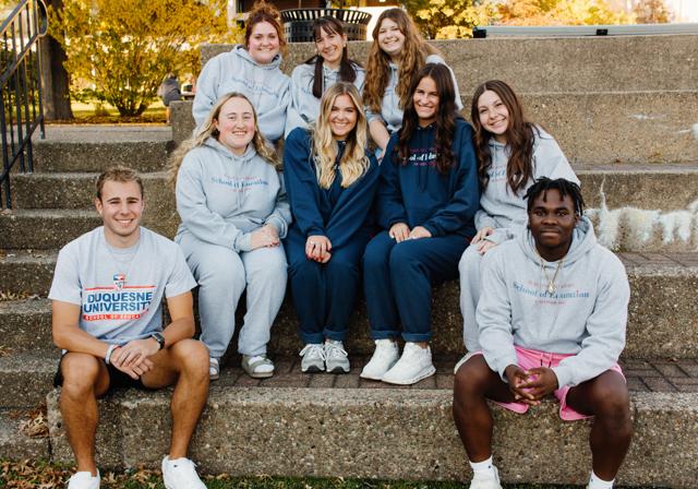 Nine School of Education scholarship students sitting on cement stadium steps in triangle formation with fall trees in background.
