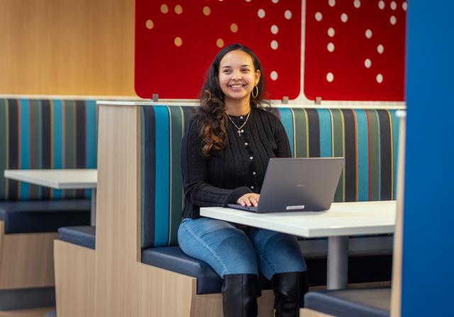 Student at laptop in booth seating looking up to the side and smiling during the child school psychology virtual open house.