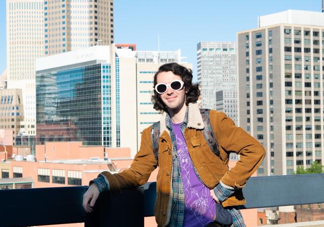 School of Education student with fun bold glasses smiling with jacket on and leaning against short wall with Pittsburgh cityscape in background