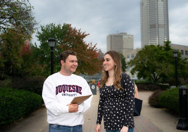 Two students smiling walking on campus with one wearing a Duquesne shirt and the cityscape in the background.