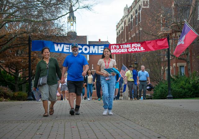 Family walking under campus banner outside that reads "Welcome It's time for bigger goals" with Duquesne flag to right, campus building and November trees in background