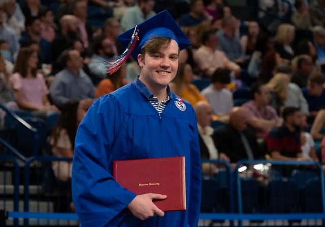 Graduate walking on commencement floor with diploma in hand and crowd behind him as he smiles and the tassel of his cap swings