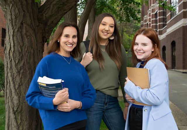 Group of three graduate students gathered smiling with books and trees and back of Canevin Hall in the background