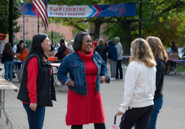 School of Education Dean, Faculty and alumni outside on campus Academic Walk and overarching sidewalk banner that reads "Homecoming and Family Weekend" with a background of Homecoming festivities 