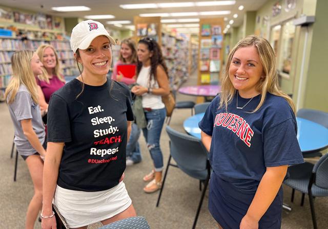 Two School of Education Student Ambassadors smiling in Curriculum Center with event attendees and colorful bookshelves behind them