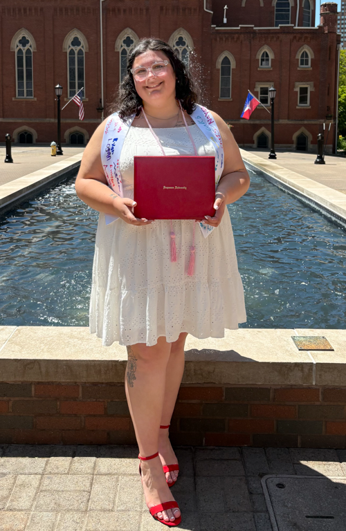 Photo of Dual Certification alum Cara Brdar holding diploma smiling outside on campus in front of waterfountain on a sunny day.