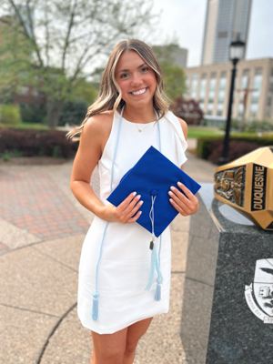 Morgan Ziolkowski smiling in graduation regalia next to Duquesne Ring monument on campus with lantern and city buildings in background
