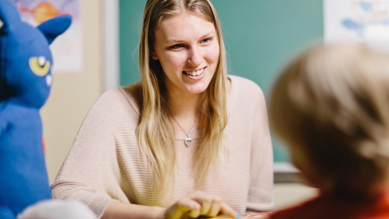 Autism Endorsement for PDE certification graduate working in clinical session with client at table pointing too book, looking at client and smiling and cat figure as teaching aid in part of session to the side.