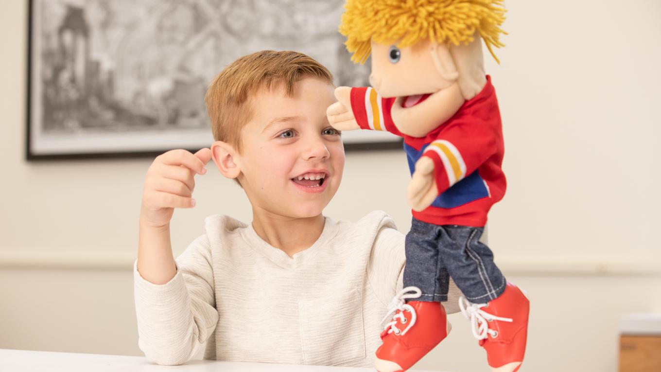 Child learning and playing with puppet that looks like him, puppet and child holding their hand up.