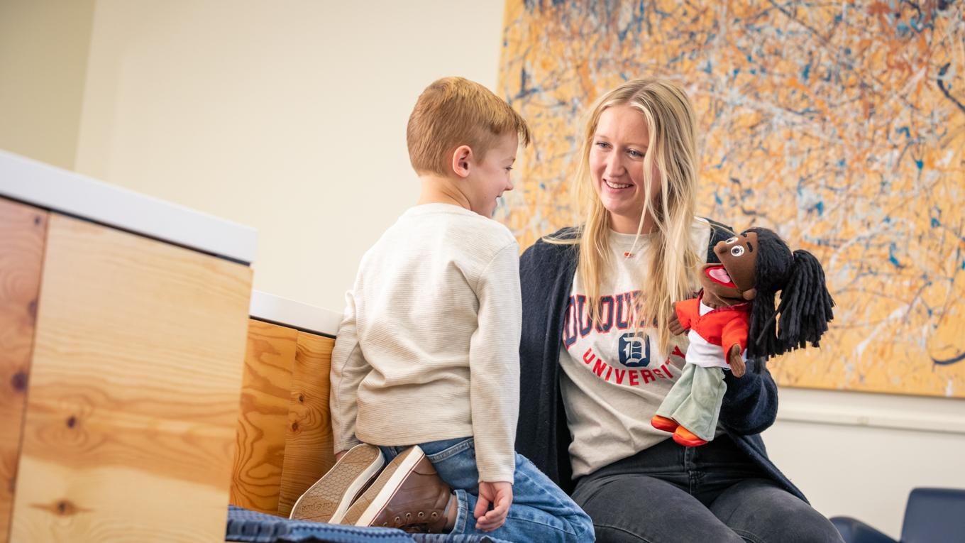 M.S. Clinical Mental Health Counseling student engaging with a young learner with a puppet on a bench in a family mental health setting. 