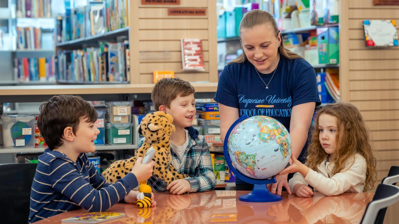 Duquesne education student teaching three young students in the library with a globe and puppet.