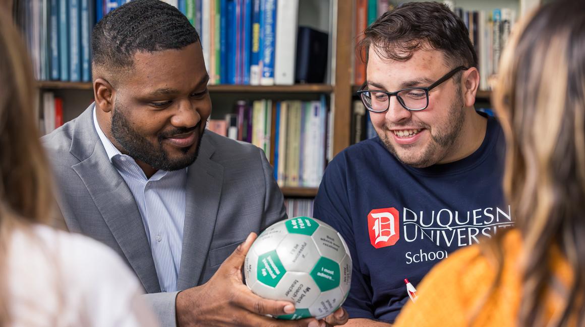 Early Education MAT Duquesne students doing mock lesson with soccer ball as faculty instructs and with book shelf in background