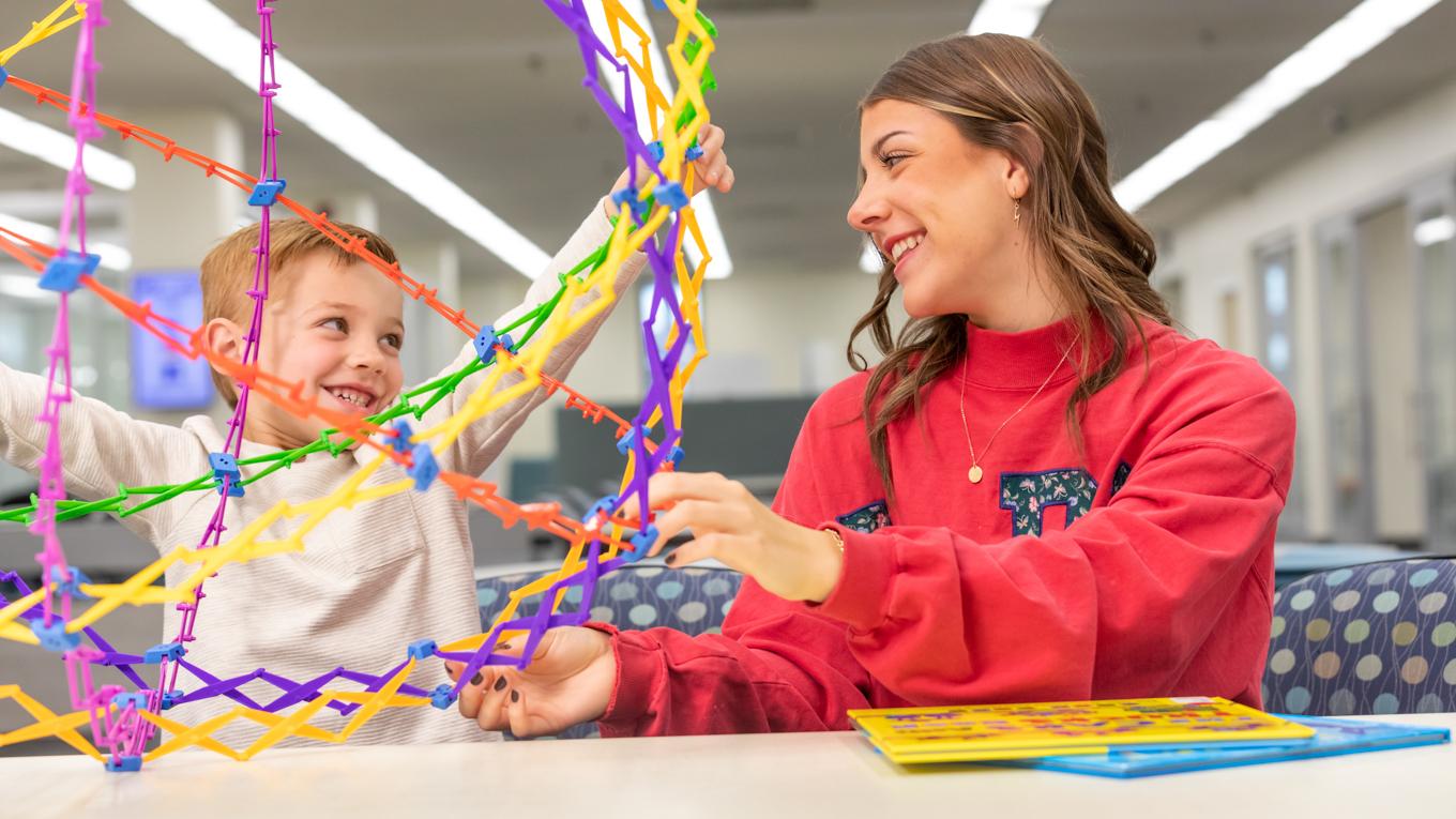 Young learner playing with geometric colorful manipulative looking and smiling at Duquesne student in library