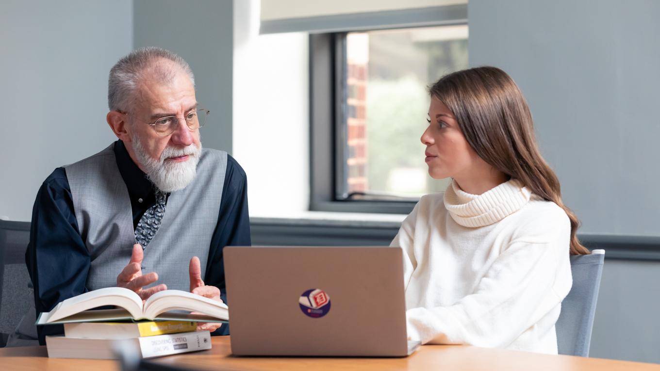 Image of professor and student talking in front of books and laptop at conference table