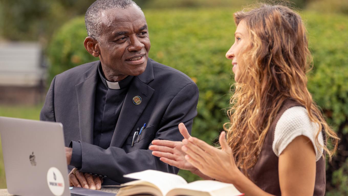 Ed.D. Educational Leadership student & priest graduate discussing research outside in Pittsburgh at table with books and laptop.