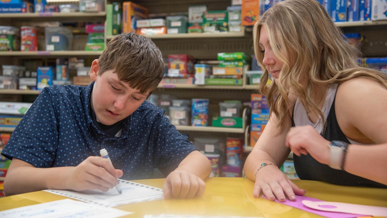 Duquesne middle level education student teacher sits at library table instructing middle school student working on lesson and colorful books in background.