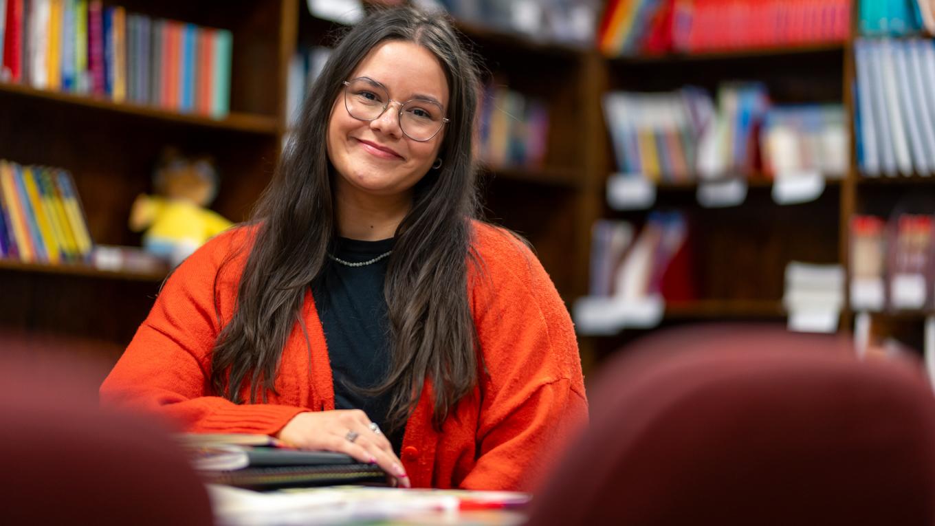 M.S.Ed. Education Administration graduate now school principal in Pennsylvania at desk smiling with notebooks surrounded by bookshelves.