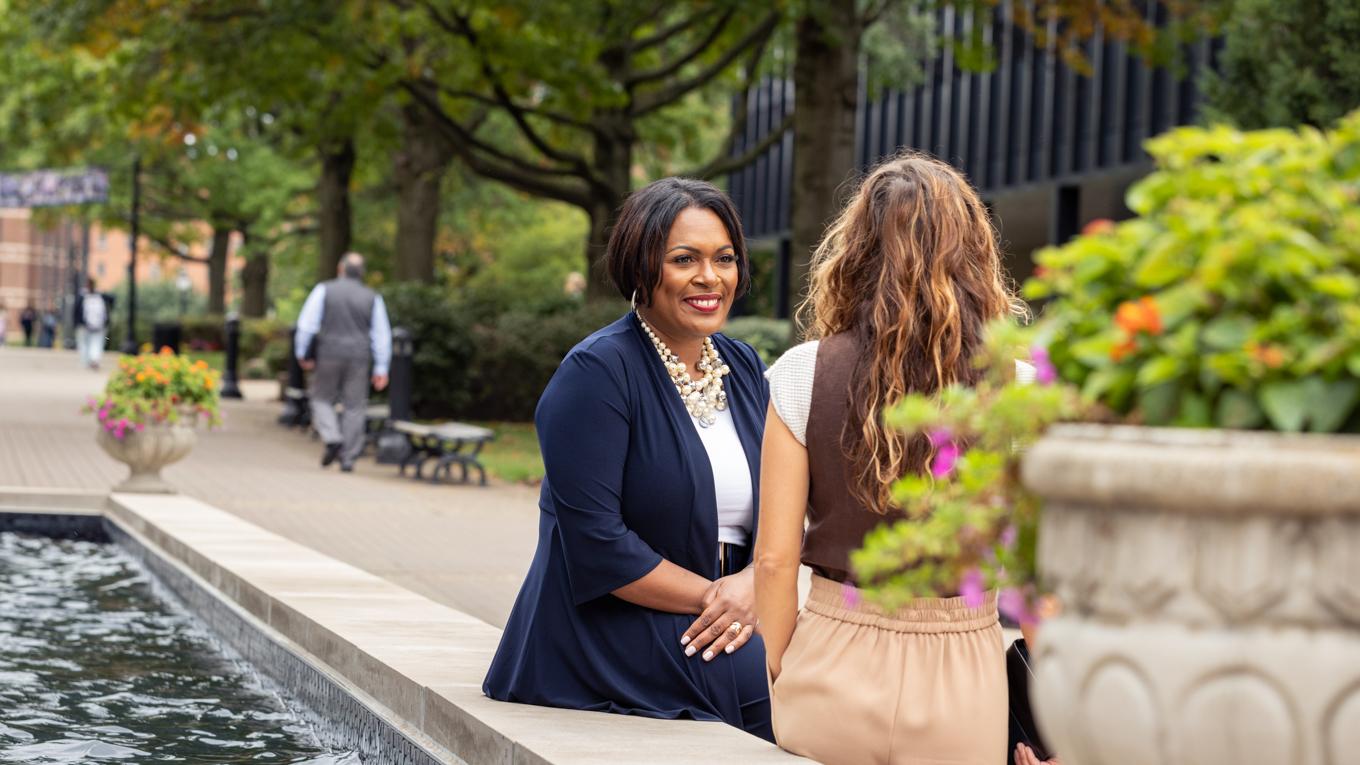 M.S.Educational Leadership master's student in discussion with Dean near water fountain outside higher education institution on summer day.