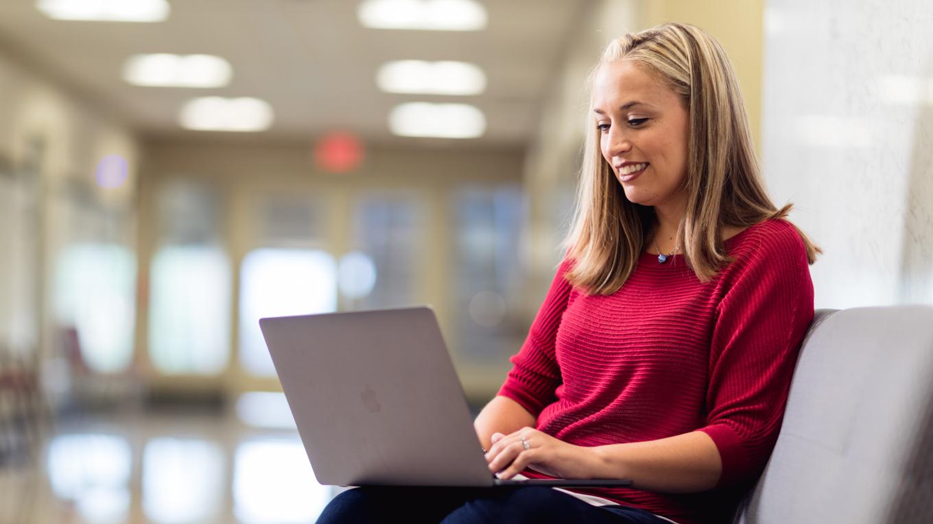 Duquense student seated in hallway bench smiling and studying on laptop