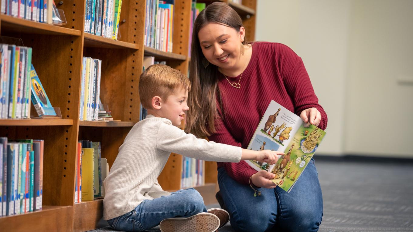 Young learning pointing at picture book Duquesne student is holding as both sit and read on the library floor